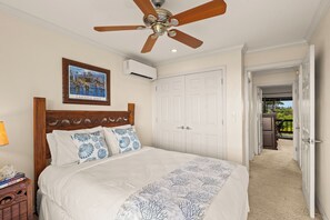 Un dormitorio con cabecera de madera, ventilador de techo, cama blanca con colcha de estampado coral y un cuadro en la pared. Se ve una puerta blanca que da a un balcón.