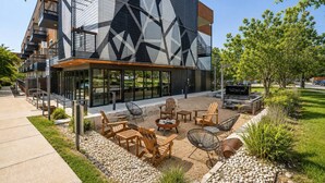 Un edificio moderno con fachada geométrica, zona de estar al aire libre con mobiliario de madera y cielo despejado.