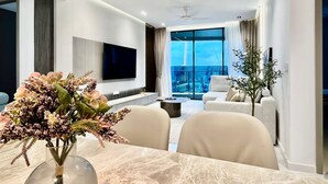 A modern living room with a flat-screen TV, a dining table with a vase of flowers, a sofa, and a view of the city. - Eaton Residences by ubook (Kuala Lumpur)