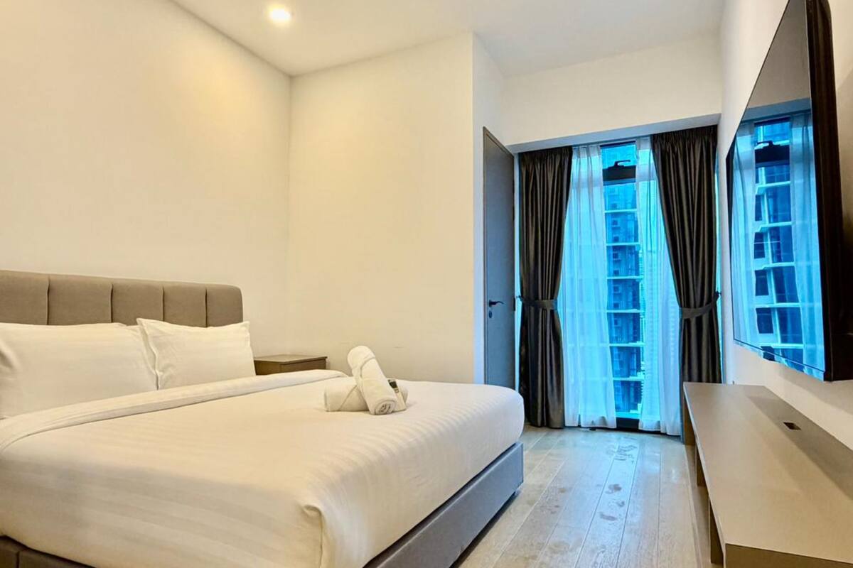 A modern hotel room with a large bed, a flat-screen TV, and a view of a cityscape through the window.