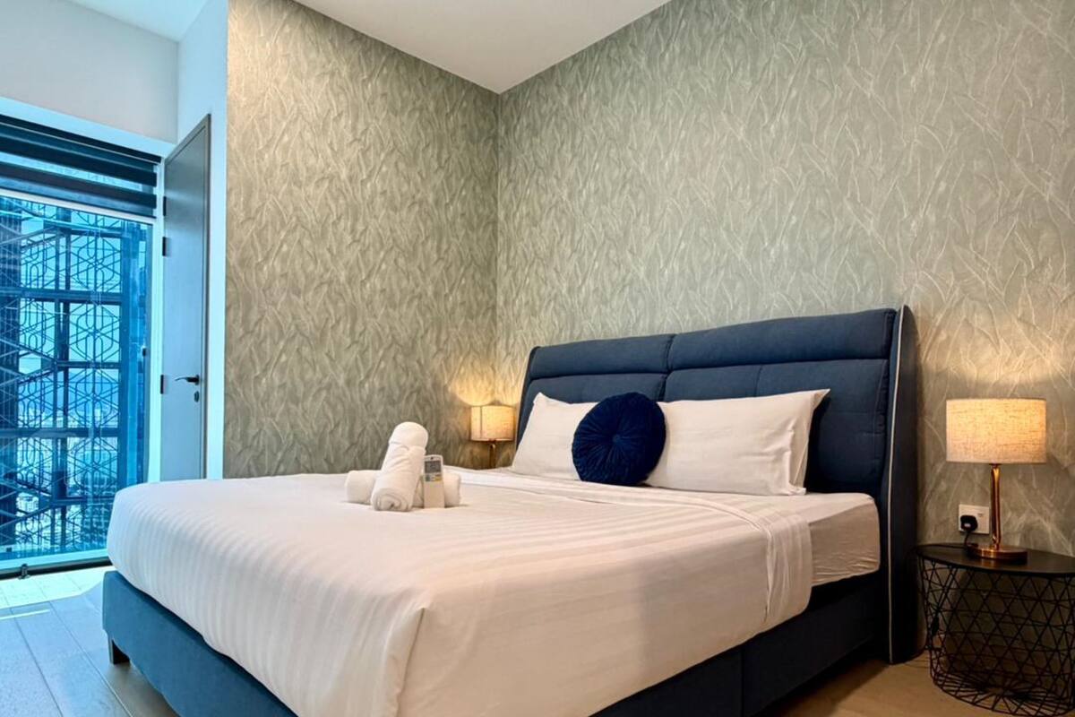 A modern hotel room with a large bed, a nightstand, a lamp, and a view of a building outside.