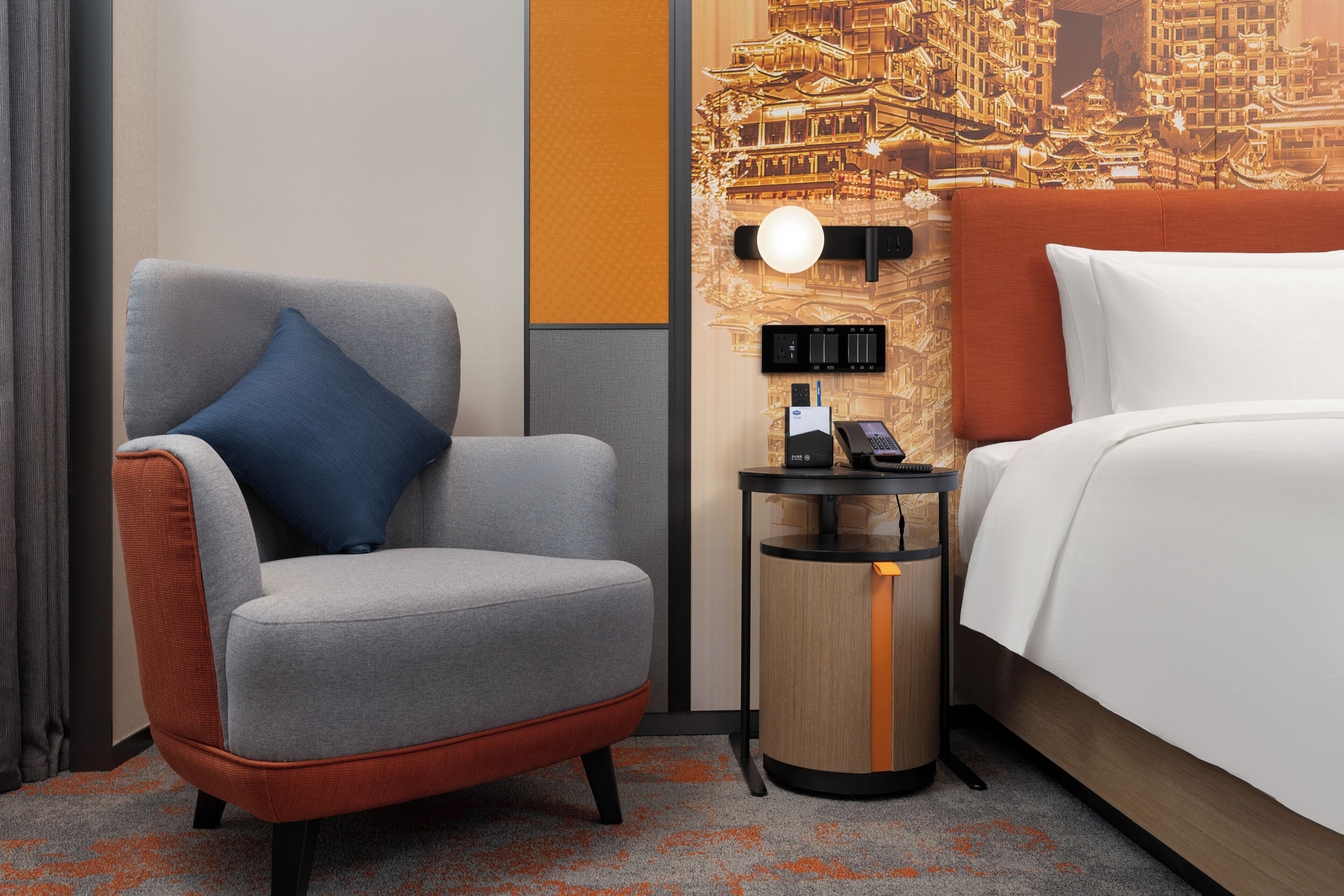 A hotel room with a grey armchair, a small round table with a phone and a lamp, and a bed with white bedding.