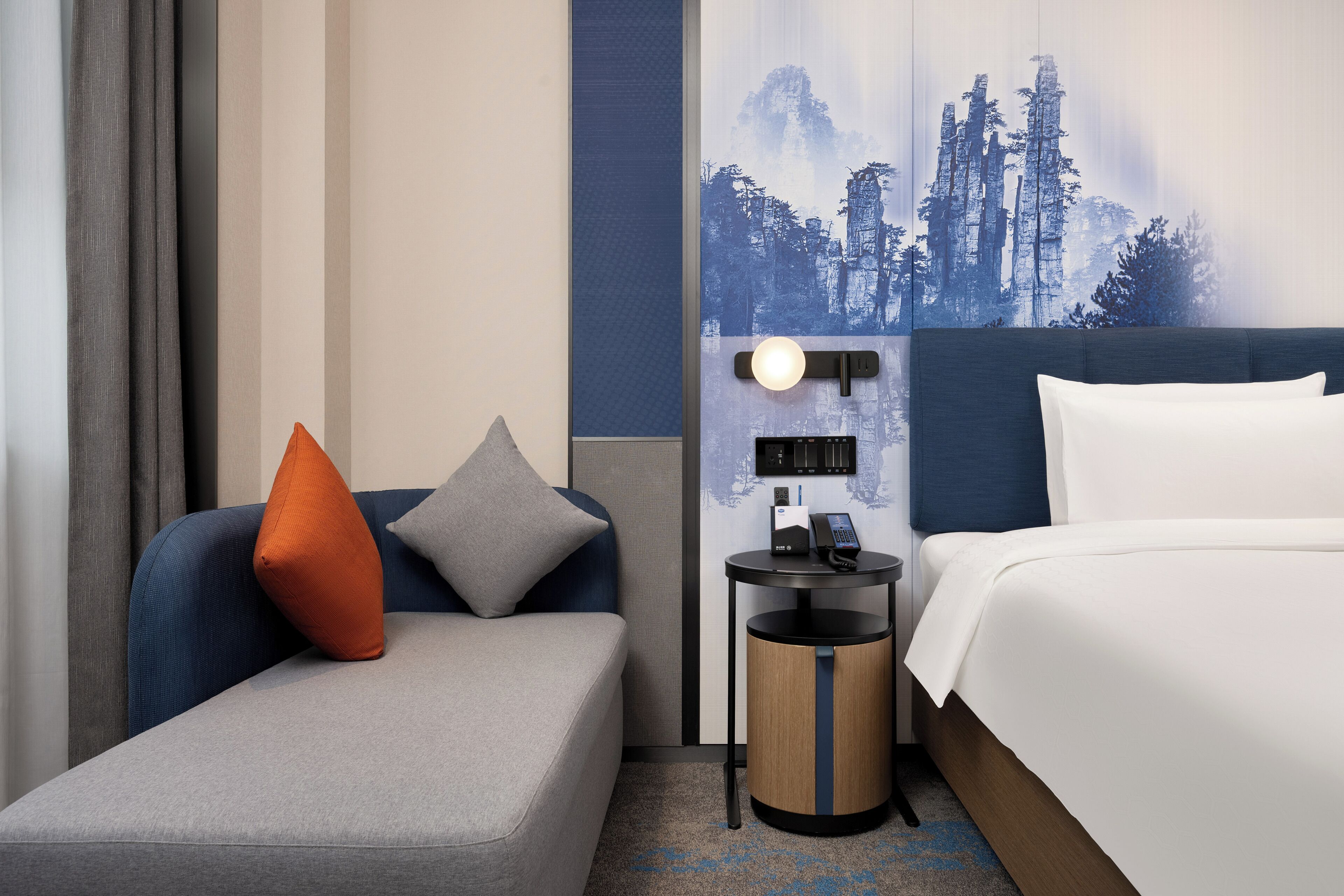 A hotel room with a bed, a small sofa, a nightstand with a lamp, and a wall-mounted phone.