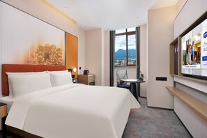 A hotel room with a large bed, a desk, a chair, a TV, and a window with a city view. - Hampton by Hilton Zhangjiajie (Zhangjiajie)