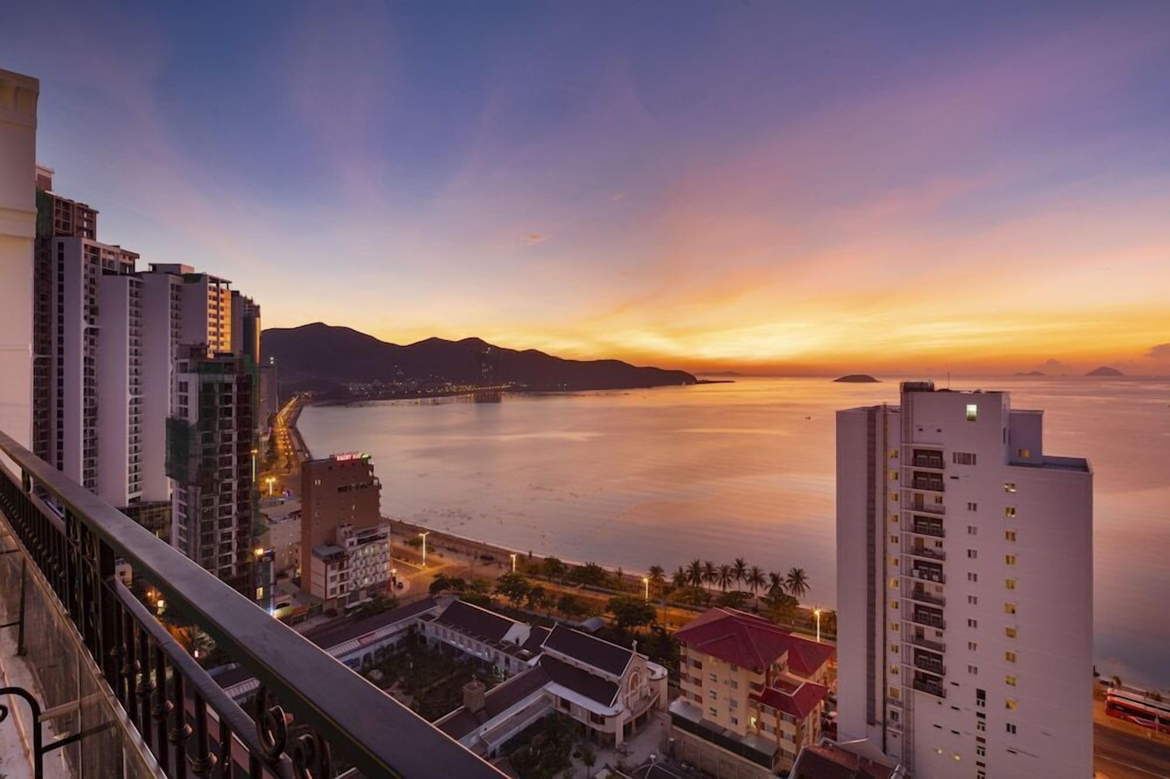 A coastal cityscape with high-rise buildings, a bridge, and a calm sea at sunset.