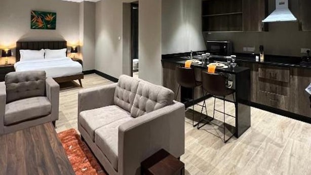 A modern hotel room with a bed, sofa, dining area, and kitchen.