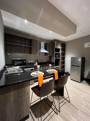 A modern kitchen with a central island, two bar stools, a built-in oven, and a refrigerator.