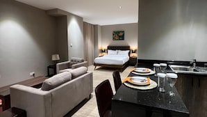 A modern hotel room with a bed, a sofa, a dining table set with plates and glasses, and a kitchen area.