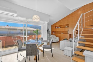 Ein modernes Wohnzimmer mit einem Glastisch, grauen Stühlen, einem weißen Sofa, einer Holztreppe und Blick auf die Stadtlandschaft.