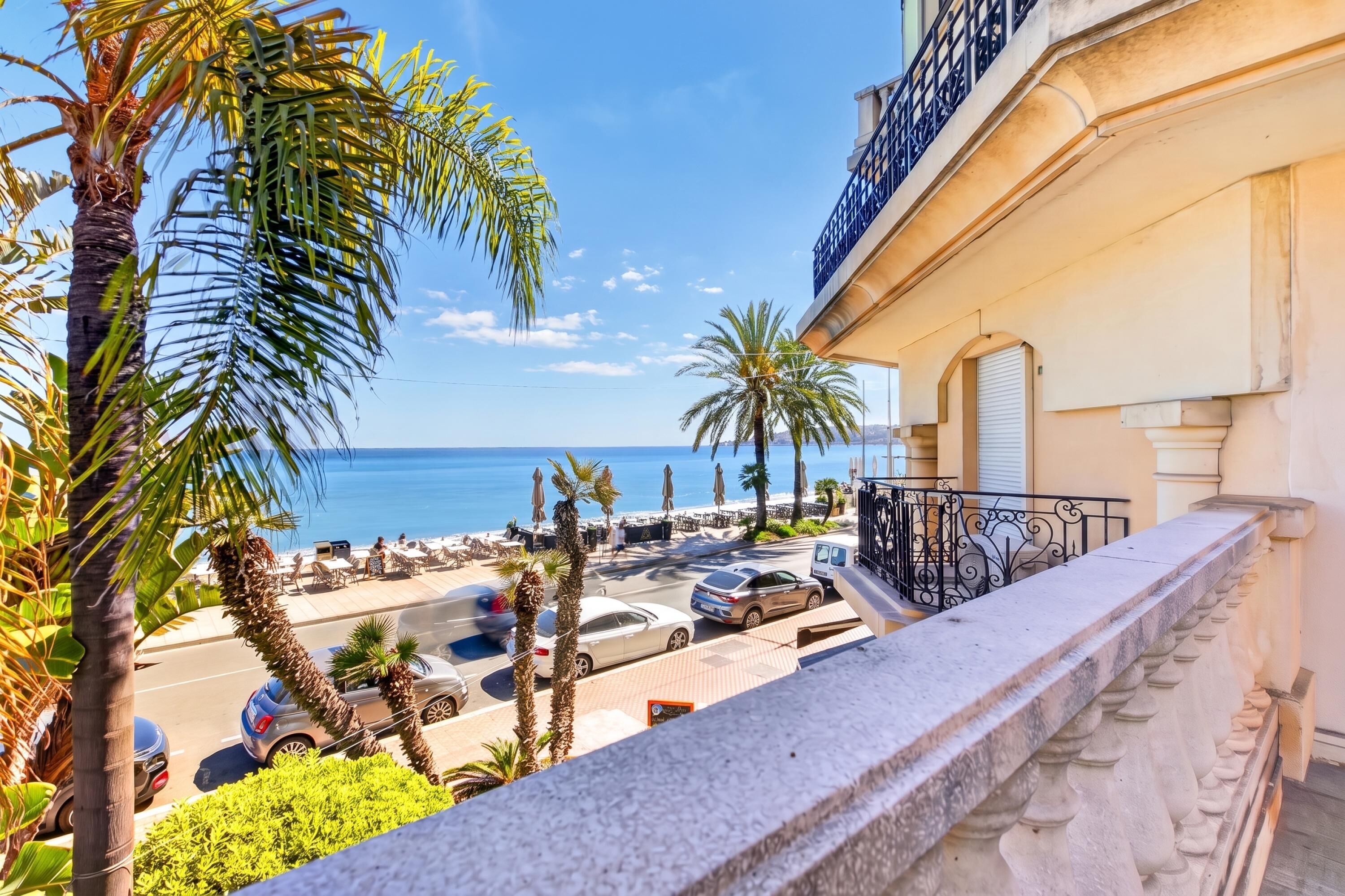 A balcony with a view of the sea, palm trees, and a street with parked cars.
