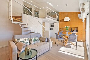 Ein zeitgemäßer Wohnraum mit einer Couch, einem Esstisch, Stühlen, einem Couchtisch, einer Treppe und einer Küchenzeile.