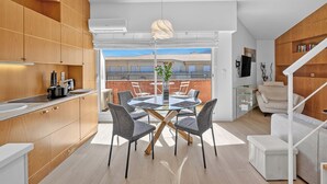 Eine moderne Küche mit Holzausstattung, ein Essbereich mit Glastisch und Stühlen sowie Blick auf ein Gebäude durch eine Glastür.