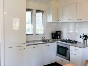 Una cocina con armarios blancos, estufa a gas, horno, cafetera y ventana con persianas.