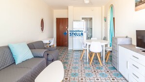 En stue med sofa, fjernsyn, spiseplads med stole og et køkken med køleskab.