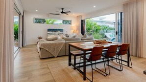 A modern living room with a sofa, a dining table with chairs, a ceiling fan, and a large window with a view of greenery. - Trinity Beach Coastal Luxe Pool Retreat – 4BR (Trinity Beach)