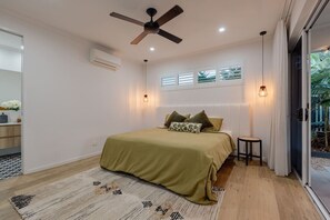 Une chambre à coucher comprenant un lit, un ventilateur de plafond, un appareil de climatisation, une table de chevet et une vue sur la verdure à l’extérieur.