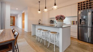A modern kitchen with a central island, bar stools, pendant lighting, and a stainless steel refrigerator. - Trinity Beach Coastal Luxe Pool Retreat – 4BR (Trinity Beach)