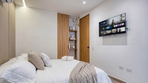 Une chambre à coucher avec un lit, des oreillers, une table de chevet, une étagère avec des livres, un téléviseur fixé au mur et une armoire en bois.
