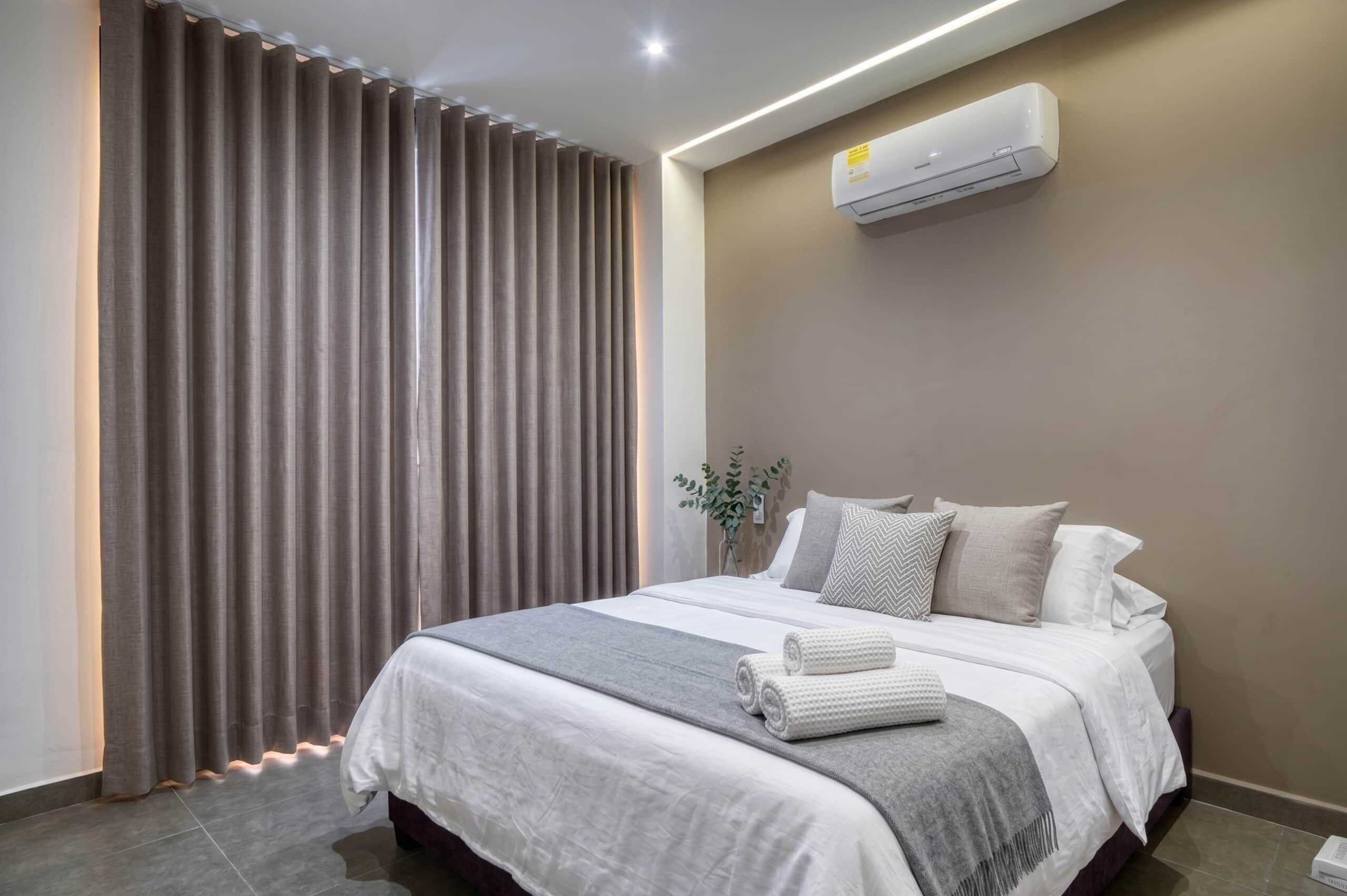 Quarto de hotel moderno com uma cama grande, cortinas cinzentas, ar condicionado fixado na parede e uma pequena planta.