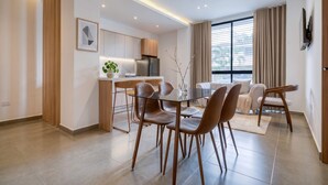 Un espace repas moderne avec une table en verre et des chaises en cuir, une cuisine dotée d’un four encastré et une grande fenêtre avec des rideaux.