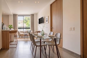 Un comedor moderno con una mesa de vidrio, sillas de cuero, un florero con ramas y un televisor en la pared.