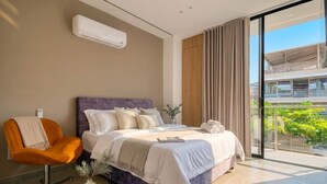 Une chambre moderne avec un lit, une chaise, une plante en pot et une vue sur un immeuble à travers une porte vitrée.
