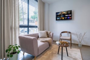 Una sala de estar con un sofá, una silla, una mesita, un televisor, una planta y una ventana con cortinas.