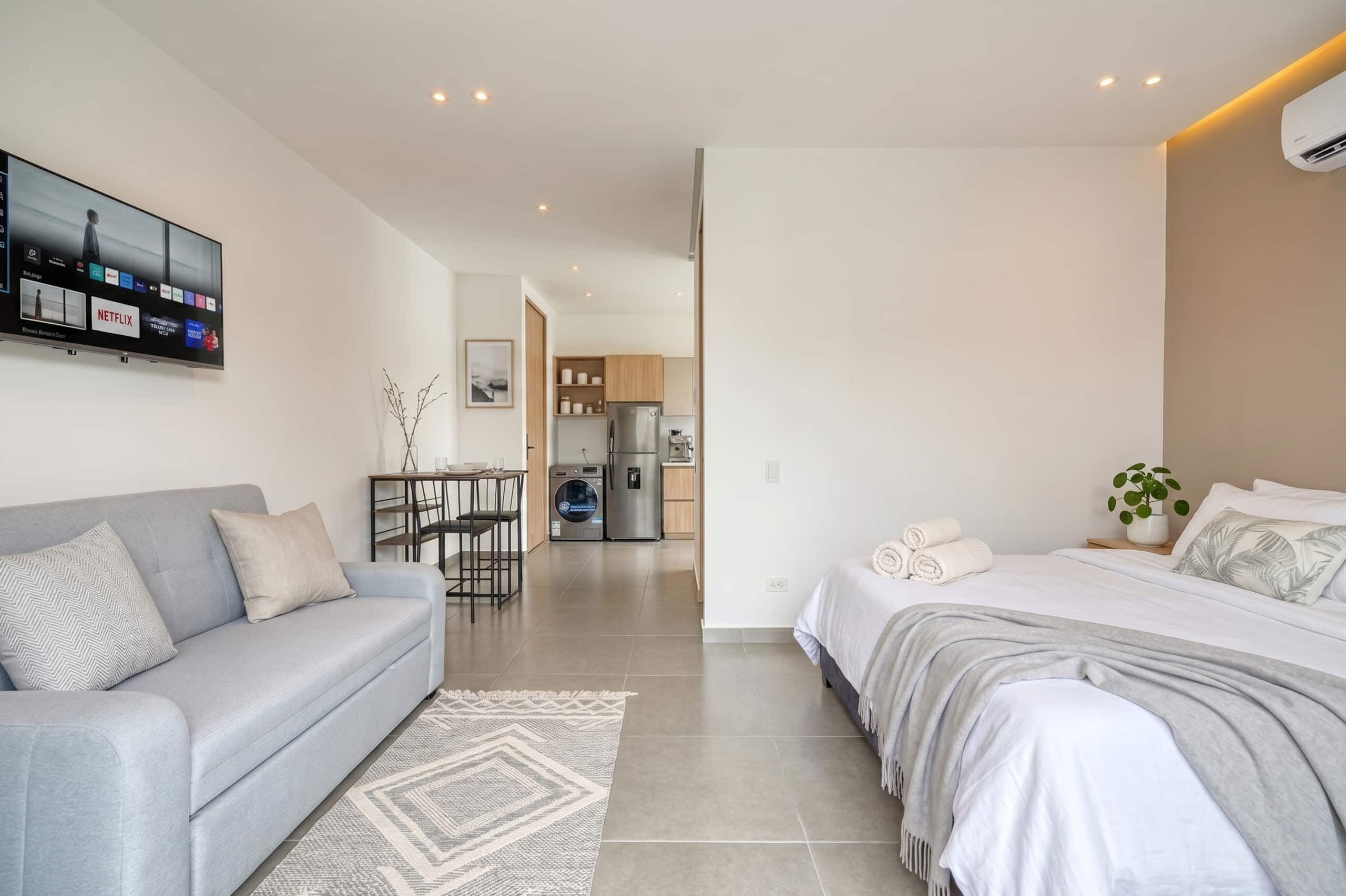 A modern hotel room with a sofa, a bed, a TV, a kitchen area, and a bathroom.