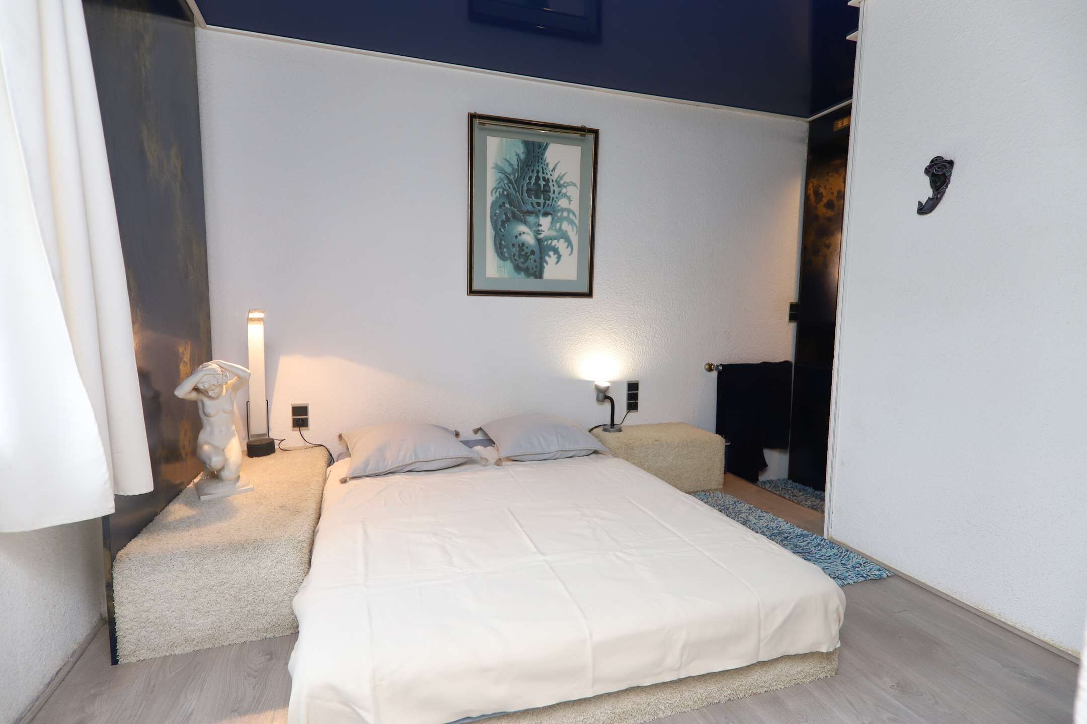 A bedroom with a bed, bedside lamps, a wall-mounted artwork, and a door with a handle.
