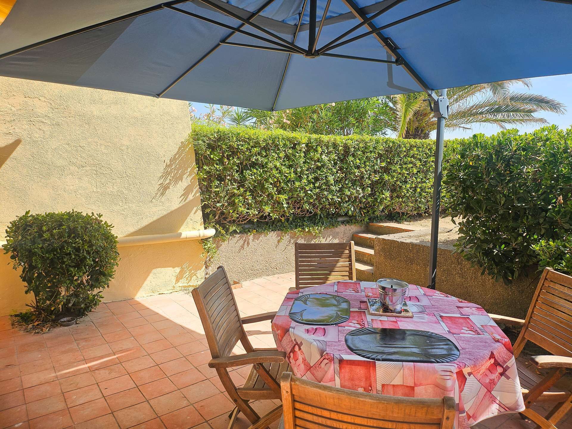 An outdoor patio with a table covered by a patterned tablecloth, surrounded by wooden chairs, under a large umbrella.