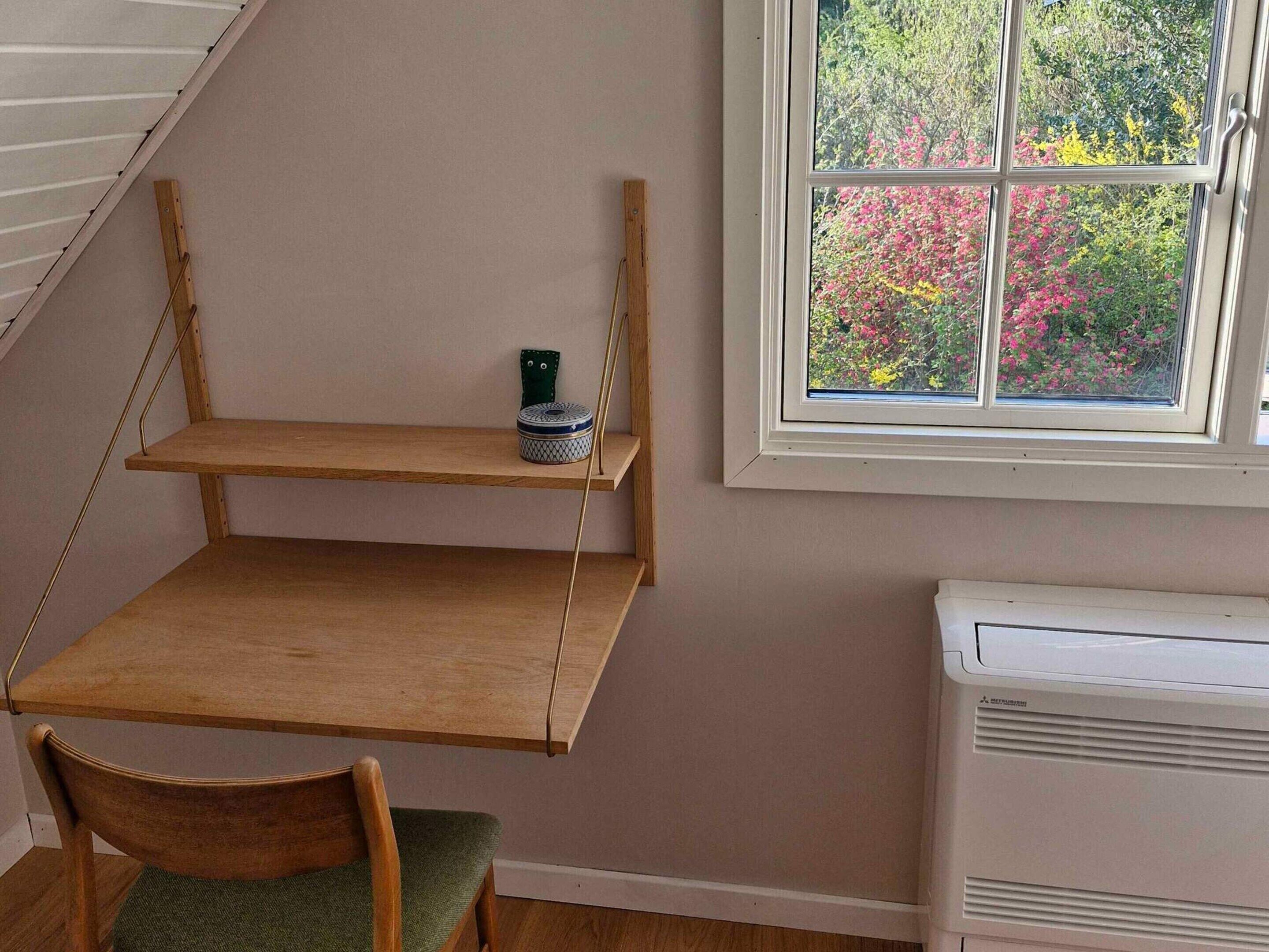 A room with a slanted ceiling, a window with a view of trees and flowers, a radiator, and a wooden chair.