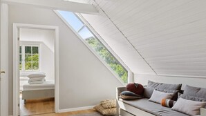 Una habitación con techo inclinado, un banco empotrado con cojines, una ventana con vistas a zonas verdes y una puerta que da a otra habitación.