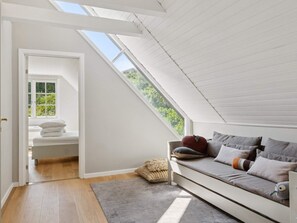 Ein Zimmer mit schräg verlaufender Decke, einer eingebauten Sitzbank mit Kissen, einem Fenster mit Blick ins Grüne und einer Tür zu einem anderen Raum.