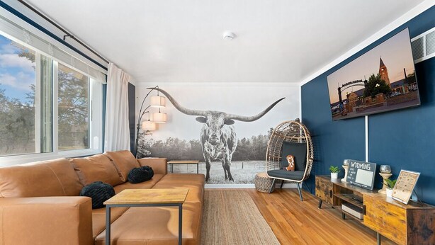 Una sala moderna con un ventanal grande, un sofá, una mesa de centro, un mural de una vaca, un televisor montado y un mueble de madera.