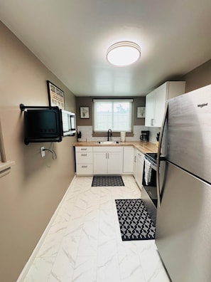 A compact kitchen with white cabinets, a built-in microwave, a stainless steel refrigerator, and a window with blinds. - The Wildflower Hideout (Cheyenne)