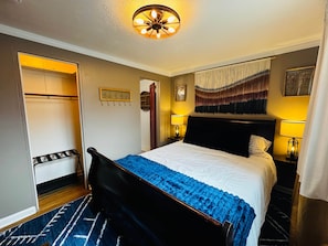 Una habitación de hotel con cama, lámparas de noche, un tapiz colgado, un armario y una lámpara de techo.