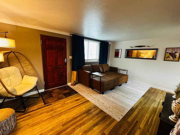 A living room with a wooden floor, a brown sofa, a yellow chair, a red door, and a window with curtains.