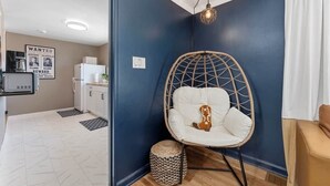 Ein Zimmer mit blauer Akzentwand, einem Hängekorbstuhl, einem kleinen Teppich und einer Küchenzeile im Hintergrund.
