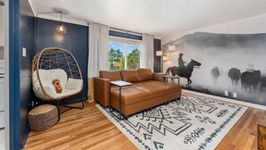 Ein modernes Wohnzimmer mit einem braunen Ledersofa, einem Korbstuhl, einem gemusterten Teppich und einem Wandbild von Pferden und Kühen.