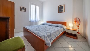 Un dormitorio con cama, mesita de noche, ventilador, una silla verde y ventana con cortinas.