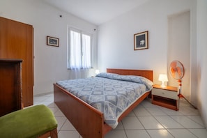 Un dormitorio con cama, mesita de noche, ventilador, una silla verde y ventana con cortinas.