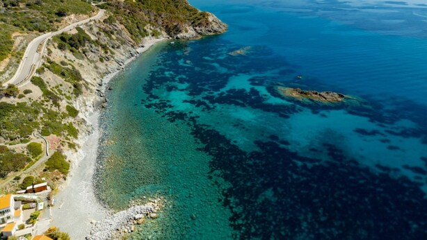 Una carretera costera serpentea a lo largo de un acantilado, conduciendo a una playa de aguas turquesas cristalinas y afloramientos rocosos.