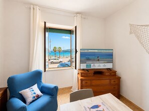 A room with a view of a beach, a television on a wooden dresser, a blue armchair, and a table with a placemat. - Le Finestre sul Porto (Portoferraio)