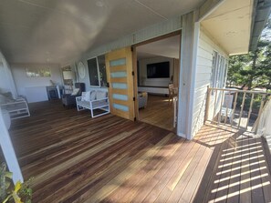 A spacious living area with a wooden deck, a sofa, a TV, and a dining area visible through an open door. - Spacious Family Retreat with Pool, Waterslide & Dual Living Minutes from Beaches (Kuluin)