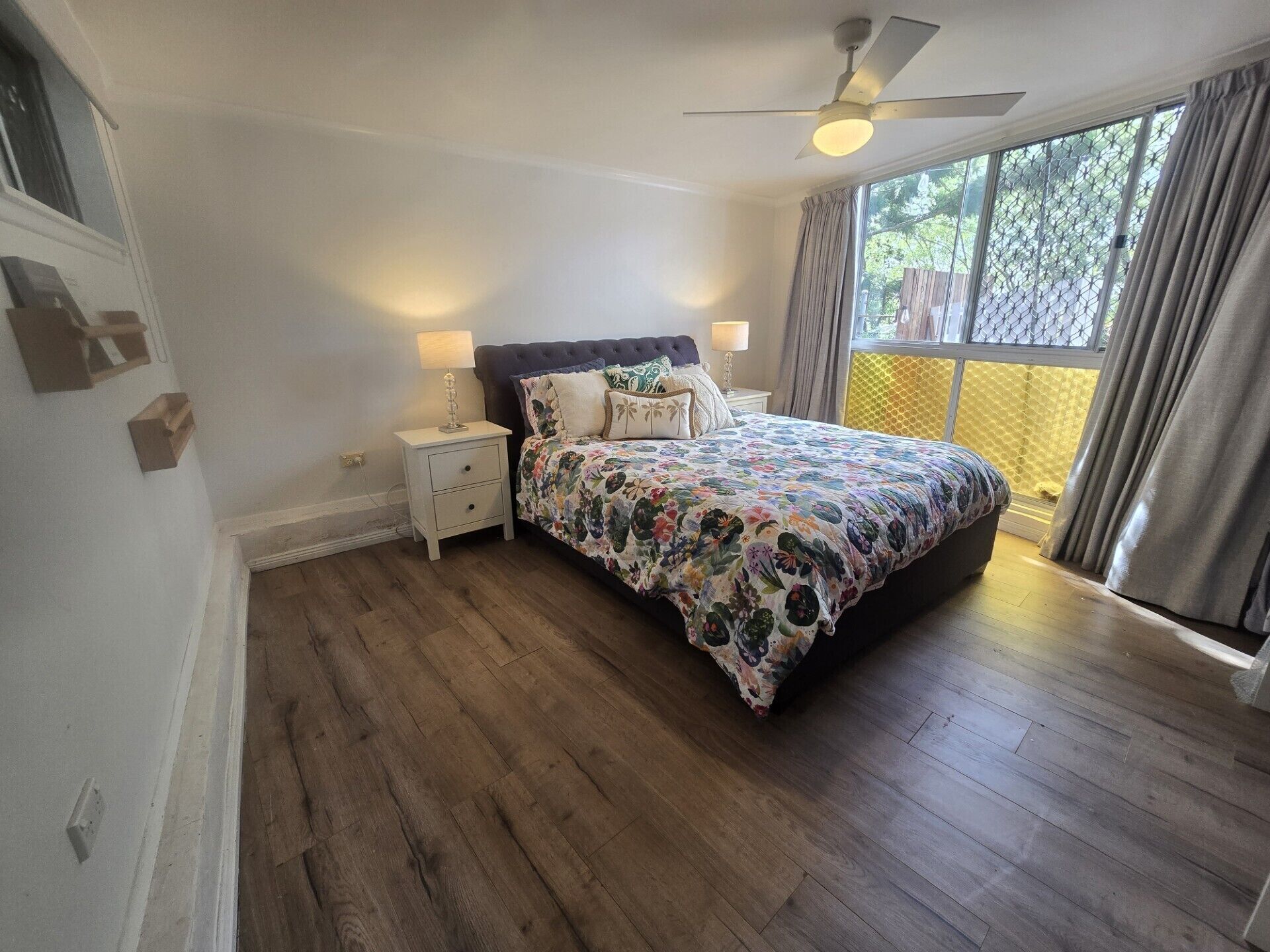 Une chambre à coucher avec un lit, des tables de chevet, un ventilateur de plafond et une fenêtre avec des rideaux.