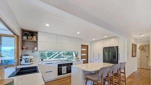 A modern kitchen with white cabinetry, a central island, and a dining area with chairs. - Spacious Family Retreat with Pool, Waterslide & Dual Living Minutes from Beaches (Kuluin)
