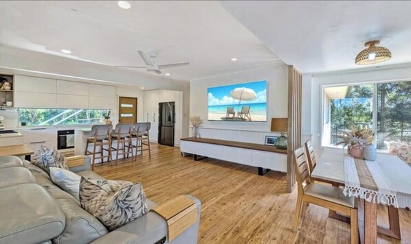 A modern kitchen with white cabinets, a dining area with a white table and chairs, and a large window with a view of greenery. - Spacious Family Retreat with Pool, Waterslide & Dual Living Minutes from Beaches (Kuluin)