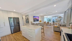 A modern kitchen with a central island, a dining area with a table and chairs, and a living space with a sofa and a TV. - Spacious Family Retreat with Pool, Waterslide & Dual Living Minutes from Beaches (Kuluin)