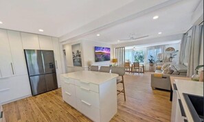 A modern kitchen with a central island, a dining area with a table and chairs, and a living space with a sofa and a TV. - Spacious Family Retreat with Pool, Waterslide & Dual Living Minutes from Beaches (Kuluin)