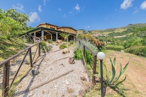 Ein Gebäude mit überdachter Terrasse, einem Kiesweg, der dorthin führt, und einer Aussicht auf eine hügelige Landschaft im Hintergrund.